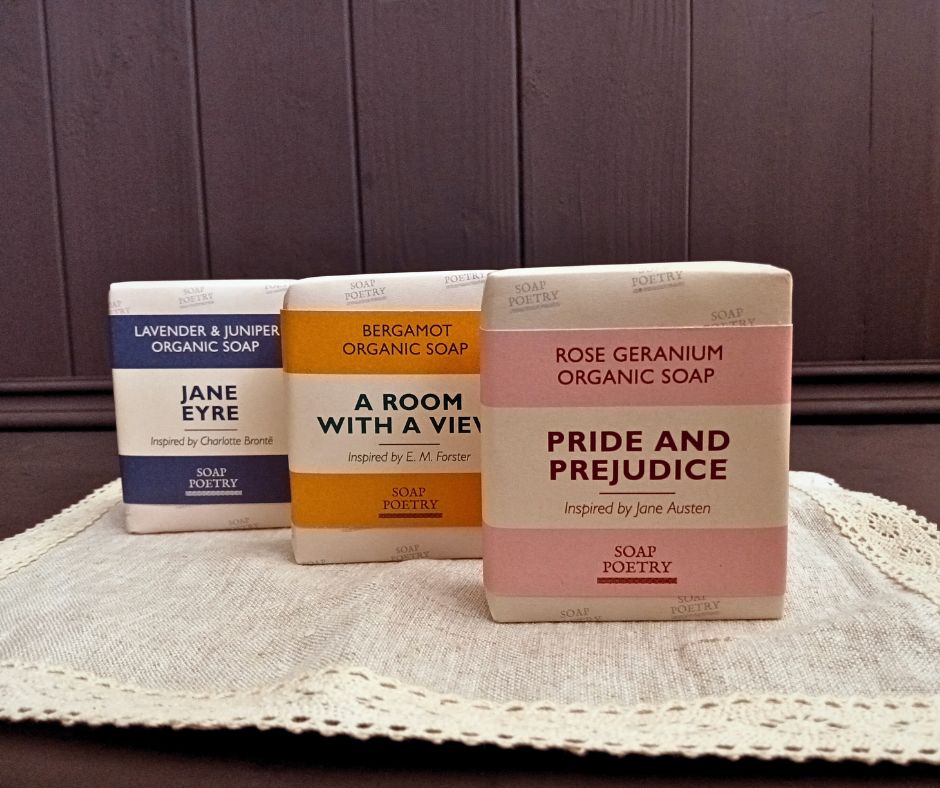 Three beautiful bars of soap with literary-themed packaging on a lace doily on a dark brown wooden base with panelling behind. .