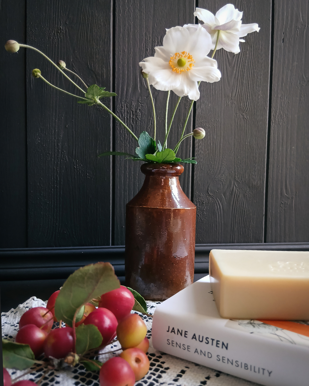 A rustic brown ceramic bottle vase holding white anemone flowers, styled on a table with a bar of soap on a Jane Austen book and red apples against a dark wood paneled wall.