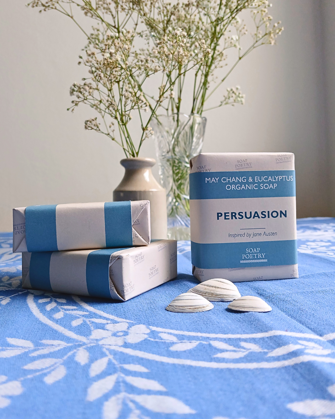 Three bars of Persuasion inspired organic soap wrapped in blue and white paper are arranged on a blue patterned tablecloth next to a glass vase with dried flowers. One bar is standing upright and labeled 'PERSUASION May Chang & Eucalyptus Organic Soap'.