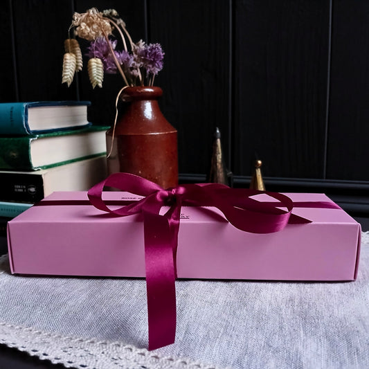 Pink gift box with a crimson ribbon on a table with books and a vase in the background.
