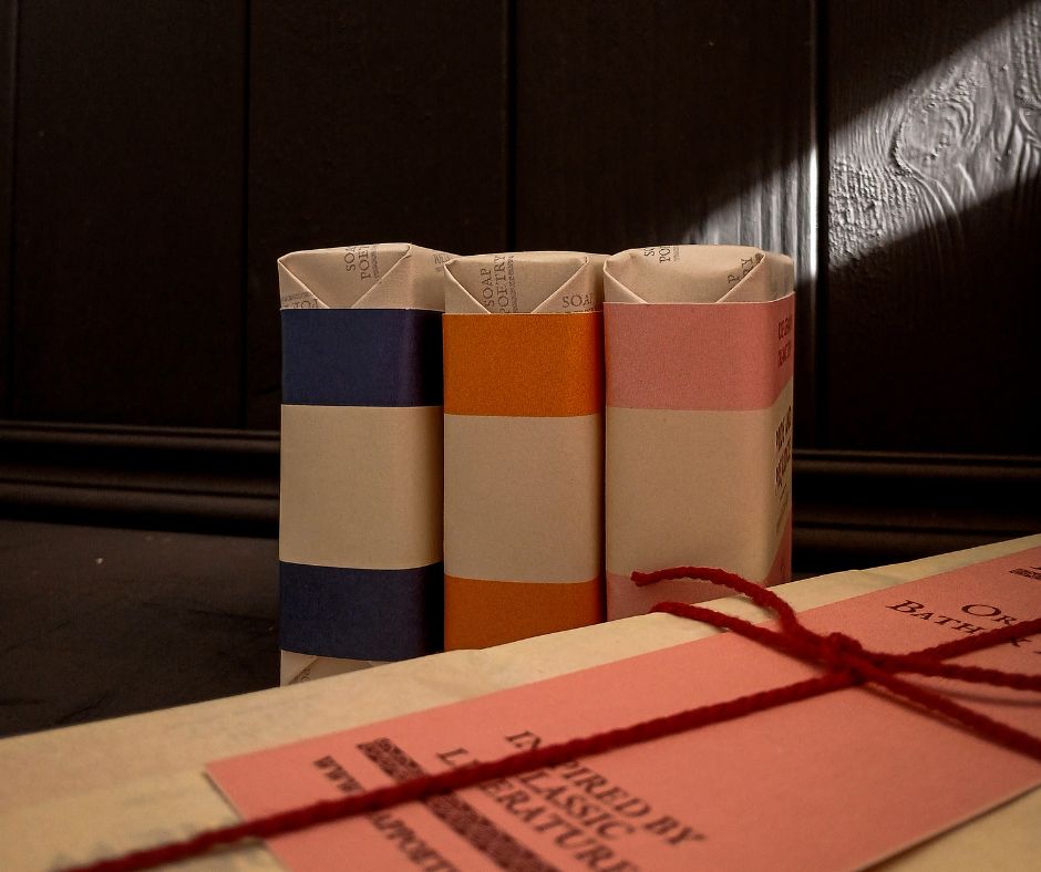 Three literary themed soaps, that wrapped in their paper resemble books with striped spines, next to a parcel with red string tied around it.