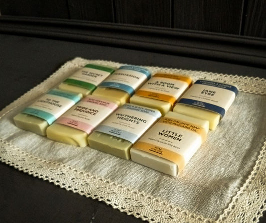 Assorted book-themed mini organic soap bars on a lace-edged linen doily with a dark panelled wooden background. 