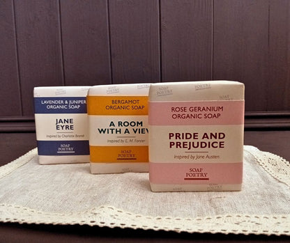 Three beautiful bars of soap with literary-themed packaging on a lace doily on a dark brown wooden base with panelling behind. .