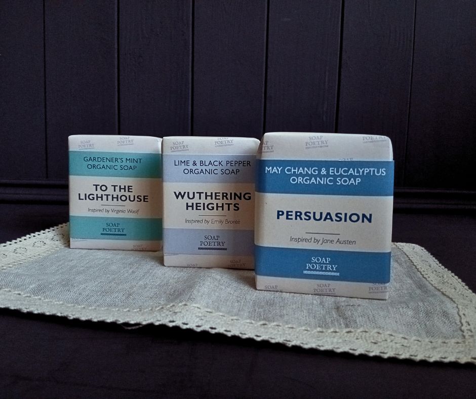 Three bars of organic artisan soap with literary-themed packaging on a textured surface. 