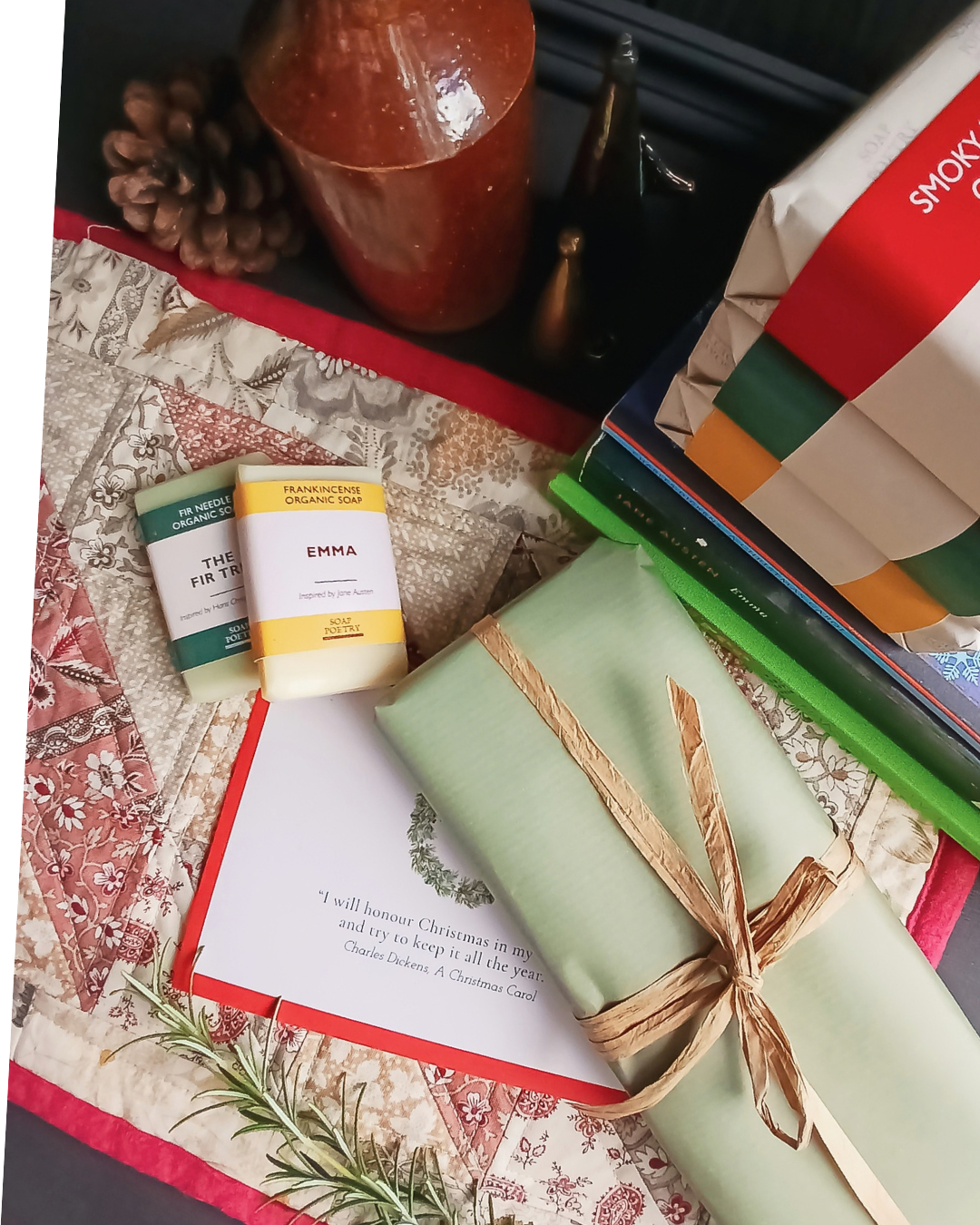 A styled flat-lay of a gift set featuring a sage green wrapped book tied with a twine bow, a small yellow and white candle labeled Emma, a small green and white soap labeled The Fir Tree, and A Christmas Carol themed card on a quilted textile background.
