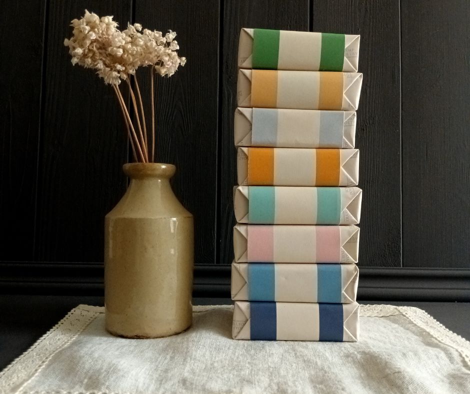 Stack of colourful wrapped soaps next to a vase with dried flowers on a linen mat with dark wood background.