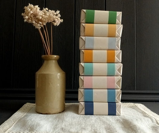 Stack of colourful wrapped soaps next to a vase with dried flowers on a linen mat with dark wood background.
