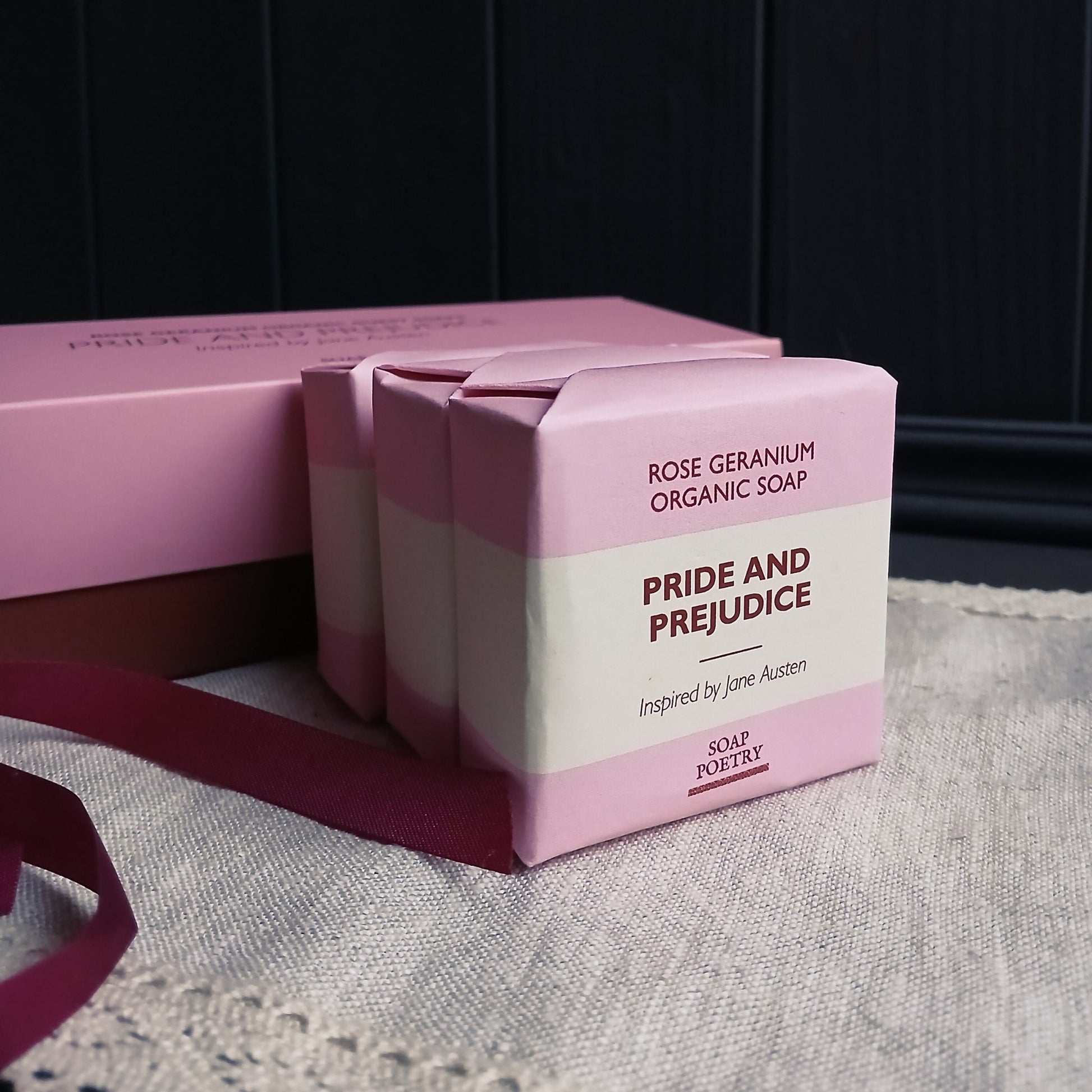 Three bars of 'Pride and Prejudice' soap beside gift box and ribbon on a linen mat. 