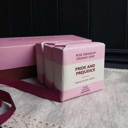 Three bars of 'Pride and Prejudice' soap beside gift box and ribbon on a linen mat. 