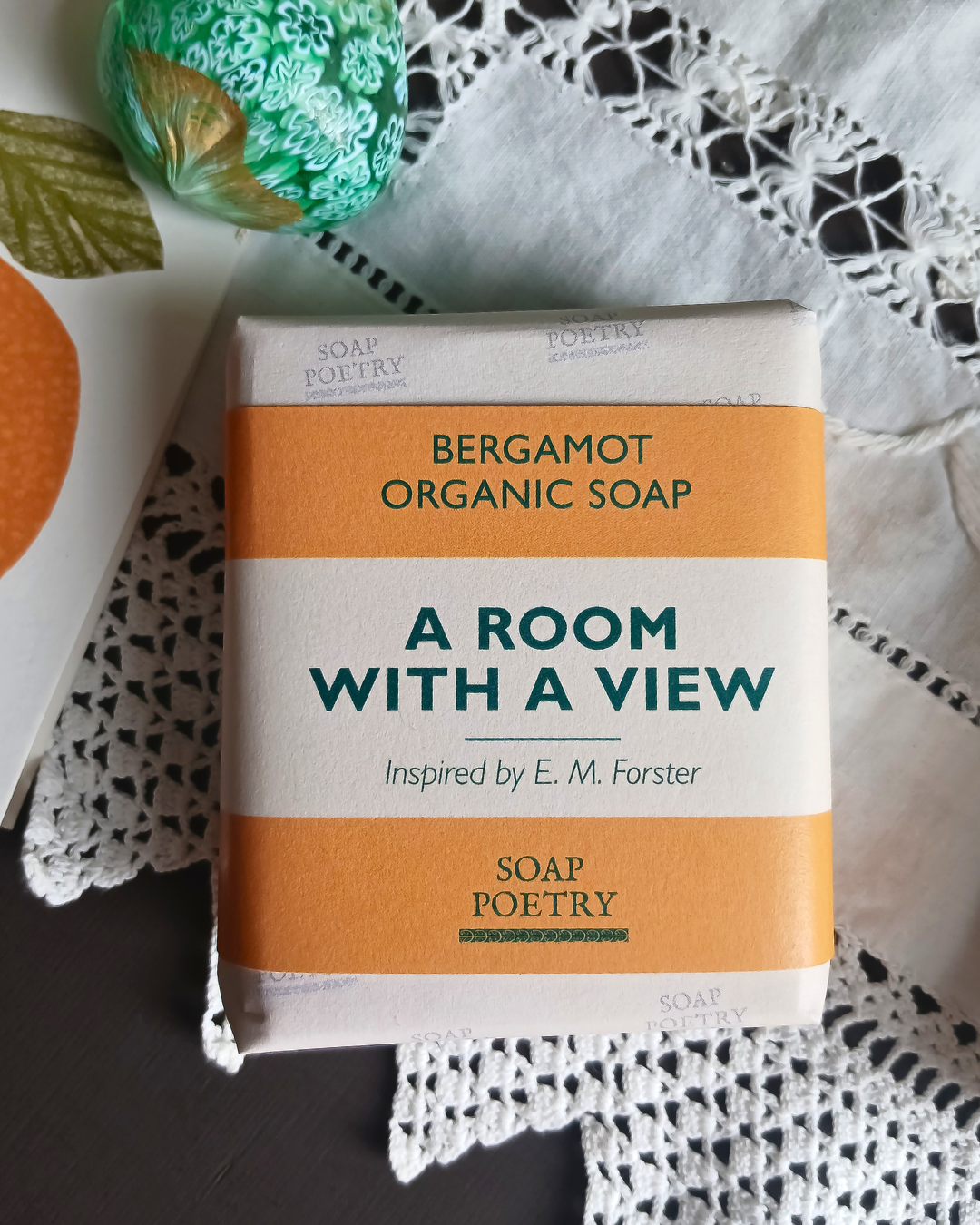 A rectangular bar of soap labeled 'BERGAMOT ORGANIC SOAP' and 'A ROOM WITH A VIEW' by Soap Poetry, wrapped in orange and off-white paper, styled on a white lace cloth with a green patterned Murano glass ornament nearby.