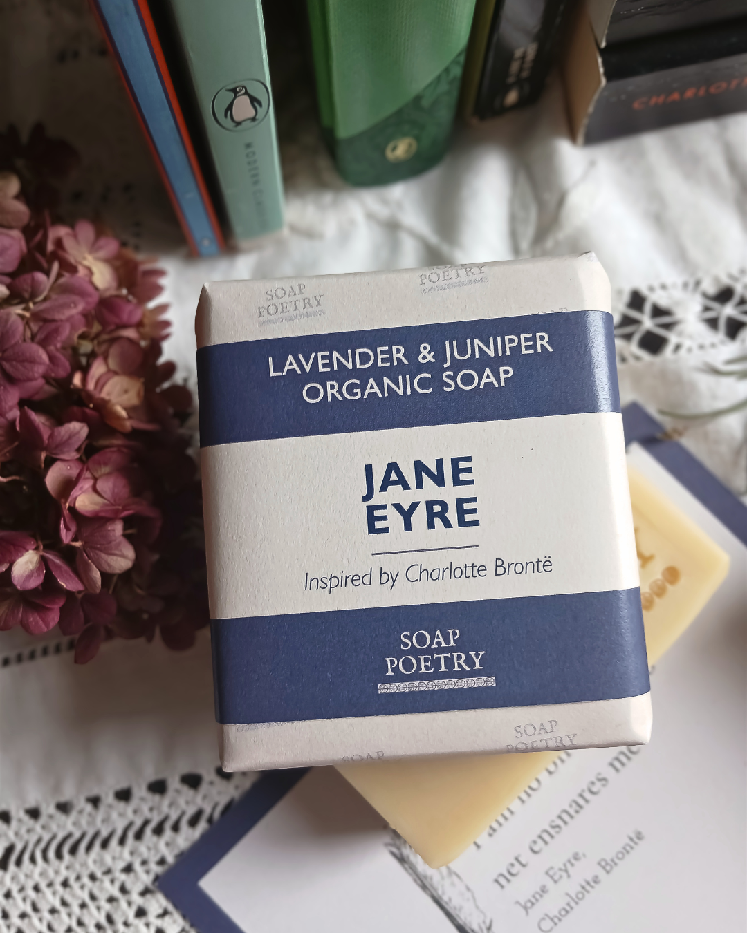 A rectangular bar of Jane Eyre Lavender & Juniper Organic Soap by Soap Poetry, wrapped in white paper with deep purple  bands, resting on a white lace cloth with books and dried flowers in the background.
