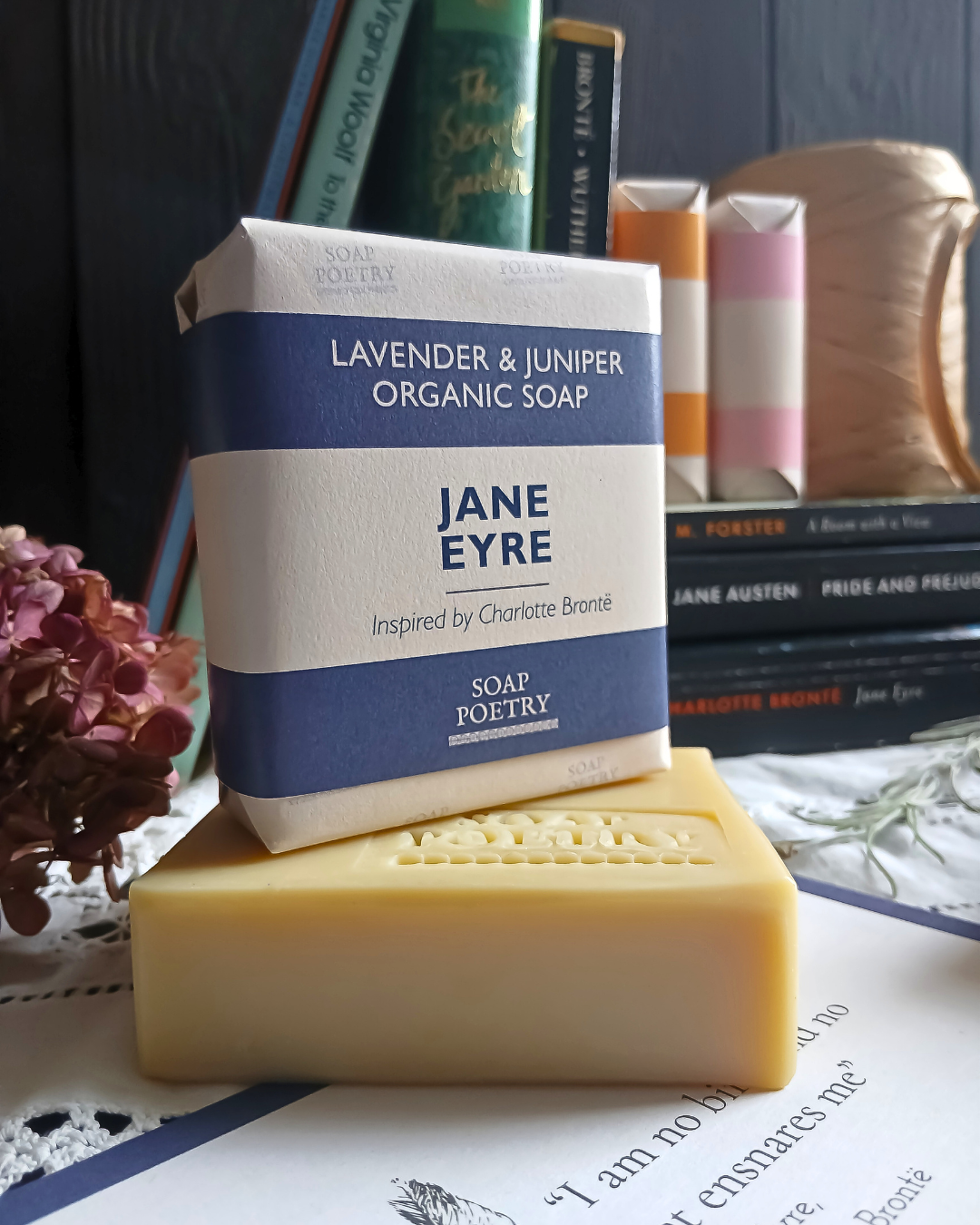 A bar of organic soap - Jane Eyre Soap Poetry - stacked on top of an unwrapped bar, styled with books and dried flowers in the background.