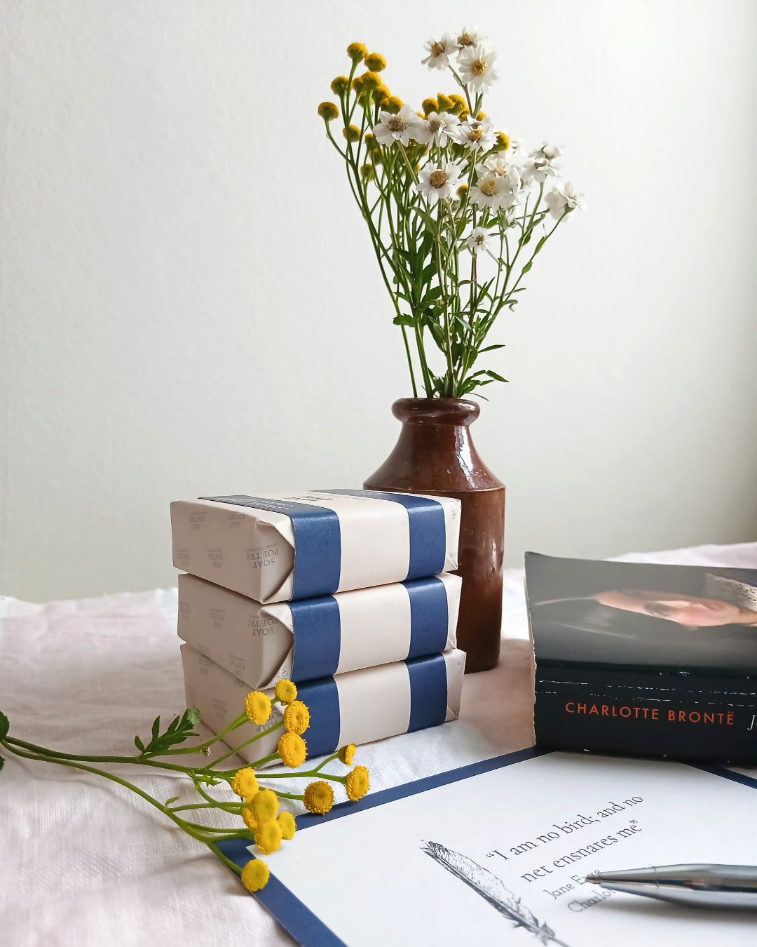 A stack of Jane Eyre Organic Soaps with a postcard and posey of flowers in a vase on a linen table cloth form cabbages and roses