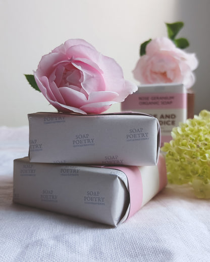 Two wrapped bars of Soap Poetry Organic Soap stacked on a white fabric surface, with a large pink rose resting on top and a second rose and soap bar blurred in the background.